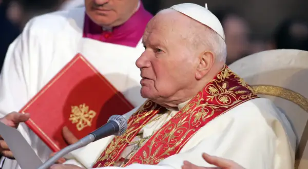Papież Jan Paweł II w białej szacie z ozdobnym kołnierzem, przed mikrofonem. Wierni odmawiają modlitwę do Jana Pawła II o uzdrowienie z raka.