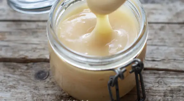 Un cucchiaio preleva il cremoso latte condensato da un barattolo di vetro su un tavolo di legno rustico.