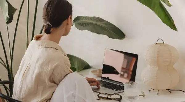 Kvinna arbetar vid laptop, omgiven av gröna växter. Kanske hon söker verktyg för att hantera kbt stress.
