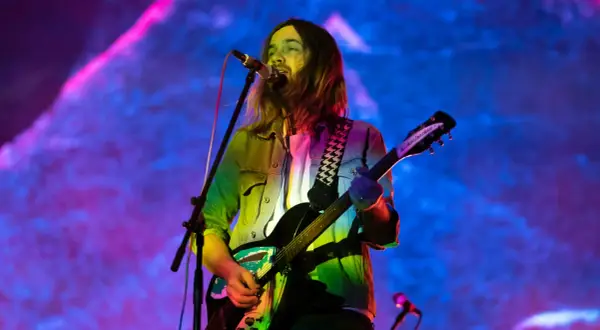 Kevin Parker z Tame Impala gra na gitarze podczas koncertu, w tle kolorowe światła.