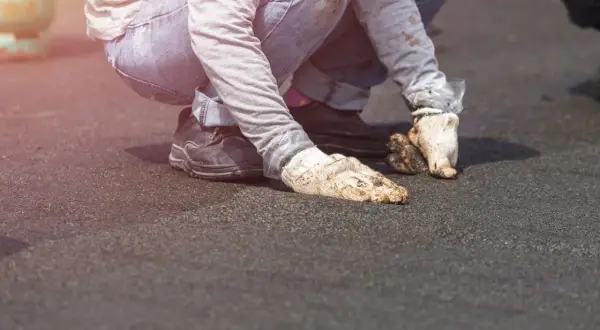 Pracownicy w brudnych rękawicach i jeansach wykonują naprawę dachu z papy, klęcząc na ciemnej powierzchni.