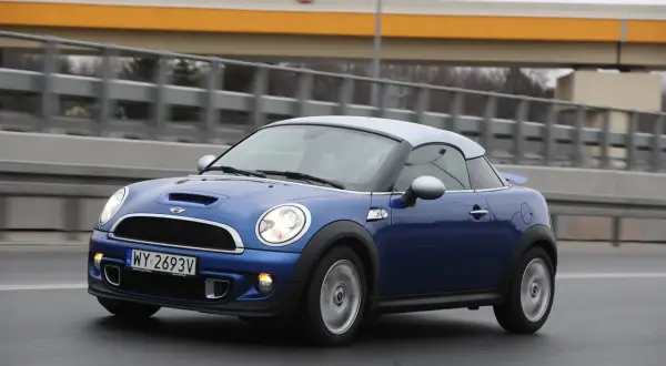 Niebieski Mini R58 Cooper S z szarym dachem pędzi po drodze, za nim widać most.