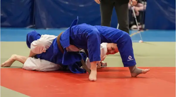 Två judokas i blå och vita dräkter kämpar på en röd matta. En av dem, med ett leende, utför en rörelse inspirerad av Jigoro Kano.