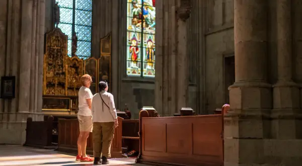 Zwei Besucher bestaunen die bunten Glasfenster im Kölner Dom. Sonnenlicht fällt durch die Fenster und malt bunte Muster auf den Boden.