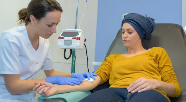 Eine Krankenschwester bereitet eine Infusion vor. Die Patientin, die eine Kopfbedeckung trägt, erhält die Behandlung, ein Zeichen dafür, dass die Chemo wirkt.