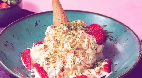 Spaghetti-Eis mit Erdbeeren und Pistazien in einer blauen Schale, garniert mit einer Eistüte.