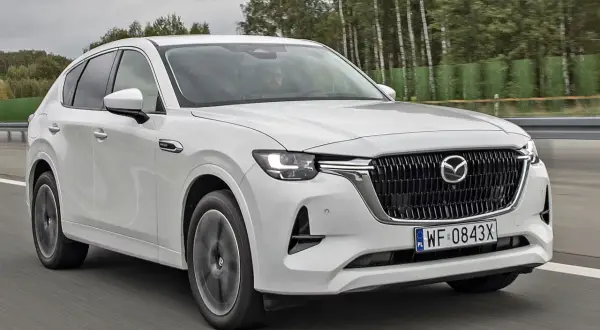 Biała Mazda CX-60 jedzie po drodze, otoczona drzewami. Sprawdź, jaka jest mazda cx 60 cena i wyposażenie.