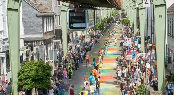 Bunte Farben auf dem Boden und eine Schwebebahn über der belebten Straße im Wuppertal Zentrum. Menschenmassen säumen die bunte Gasse.
