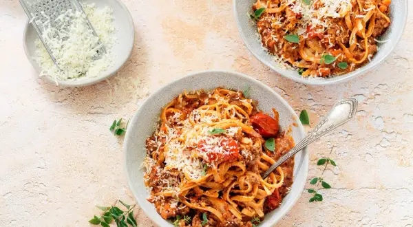 Dwa talerze spaghetti bolognese, posypane parmezanem. Obok miseczka z tartym serem i tarka. Długie gotowanie sosu to klucz do głębi smaku.