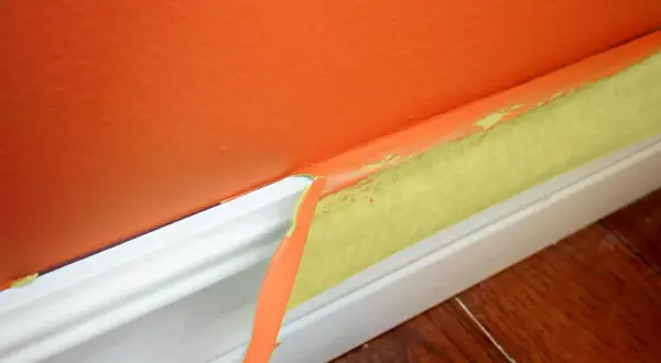 Ta bort maskeringstejp efter målning ger en ren kant. Orange vägg möter vit golvlist med gul tejp.