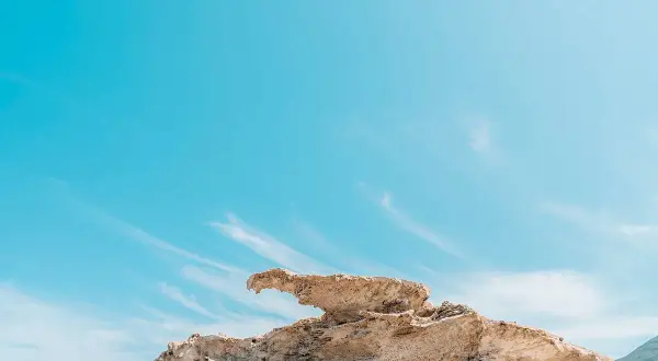 Roca erosionada con forma de ave en una playa de arcos, bajo un cielo azul intenso con nubes finas.