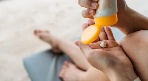 Manos aplicando crema solar caduca en la palma. Un niño descansa en una tumbona al fondo.