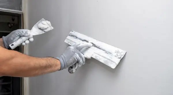 Main tenant, avec des gants, applique du plâtre sur un mur. Il utilise deux truelles pour lisser la surface, une étape essentielle pour savoir comment enlever du crépi.