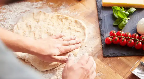 Hände formen den **pizzateig auftauen** auf einer bemehlten Holzoberfläche. Daneben liegen frische Tomaten, Mozzarella und Basilikum.