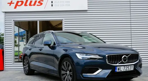 Ciemnoniebieskie Volvo V60 Hybrid z polskimi tablicami WL 7253Y przed budynkiem z logo "+plus".