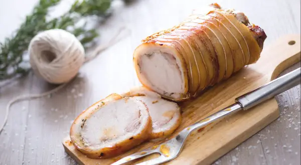 Ein aufgeschnittener, goldbraun gebratener Schweinebraten (Porchetta) auf einem Holzbrett mit Gabel und Rosmarin.