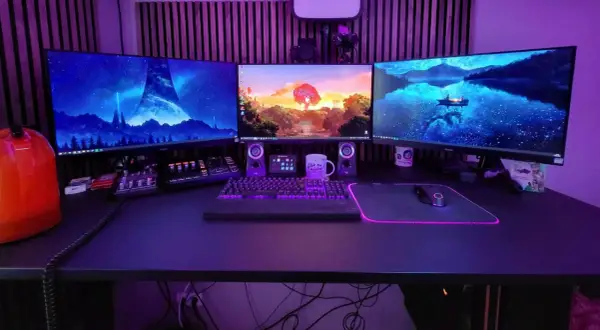 Ein episches **gaming set up** mit drei Monitoren, beleuchteter Tastatur und Maus. Im Hintergrund leuchtet lila Beleuchtung.