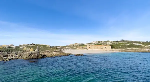 Vista de la isla de Sálvora con sus rocas, una playa de arena y un edificio de piedra bajo un cielo azul.