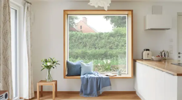 Gemütliche Fensterbank mit Kissen und Decke, ein Buch liegt offen. Das große Fenster ohne Gardinen gibt den Blick auf einen üppigen Garten frei.
