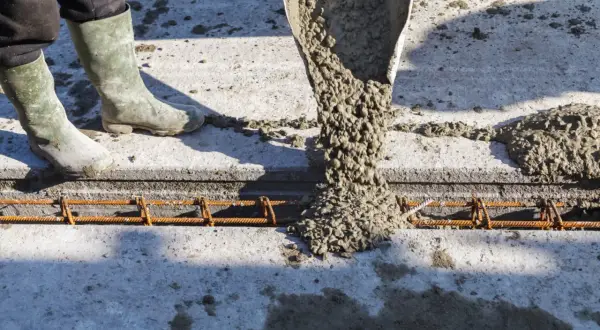 Beton wylewany z rury na zbrojenie. Pracownik w gumowcach stoi obok, nadzorując proces.