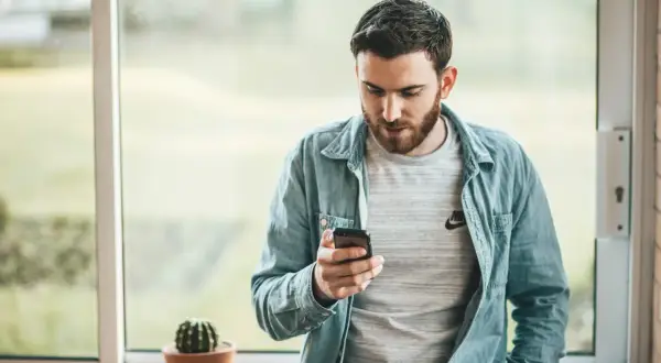 Hombre con barba mira su móvil, quizás buscando frases cuando no te responden. Un cactus en maceta decora el alféizar.