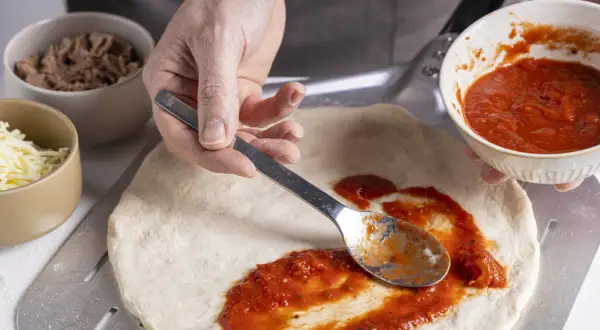 Zubereitung eines italienischen Thunfisch Pizza Rezepts: Tomatensauce wird auf den Teig gestrichen. Daneben liegen Thunfisch und Käse.
