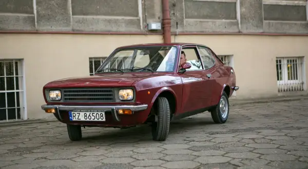 Czerwony fiat 128, klasyk z lat 70., zaparkowany na brukowanym dziedzińcu przed budynkiem.