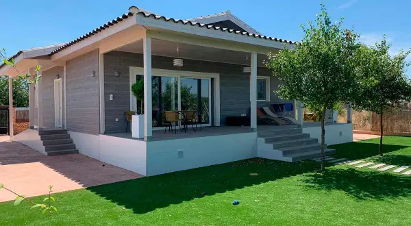 Moderna casa Canexel con porche, césped artificial y árboles frondosos bajo un cielo azul.