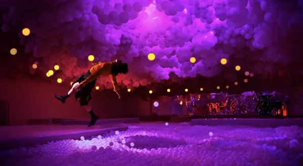 A person in a yellow shirt leaps into a ball pit at the balloon museum London, surrounded by purple clouds and yellow lights.