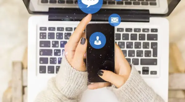 Dłonie trzymające smartfon nad klawiaturą laptopa. Ikony komunikatorów unoszą się w powietrzu. Zastanawiasz się, dlaczego na Messengerze nie widać, kiedy ktoś był aktywny?
