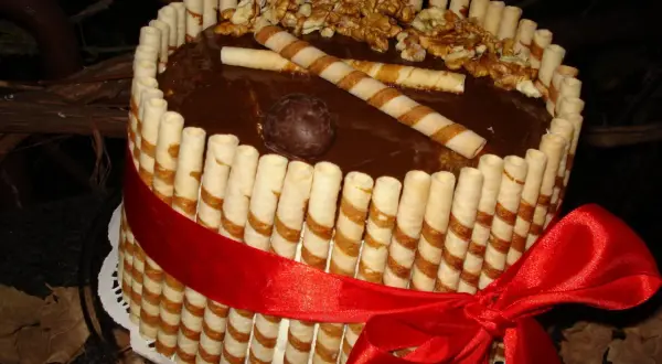 Wyjątkowy tort z rurkami waflowymi, polany czekoladą, ozdobiony orzechami i czerwoną wstążką.
