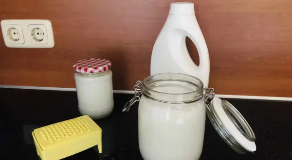 Zelf wasmiddel maken met dille en kamille. Glazen potten met wit wasmiddel, een gele rasp en een witte fles staan op het aanrecht.