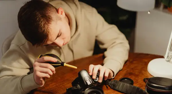 A young person carefully performs camera cleaning, using a blower to remove dust from their DSLR.