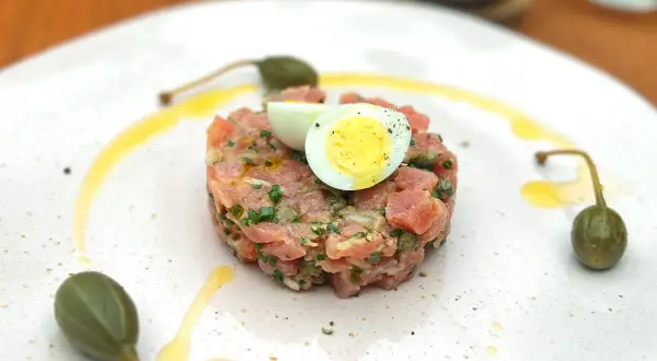 Un délicieux tartare de thon, garni d'un œuf de caille et de câpres, est servi sur une assiette blanche avec un filet d'huile d'olive.