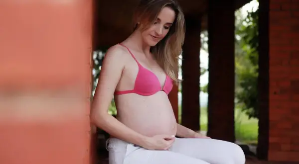 Kobieta w ciąży w różowym biustonoszu i białych spodniach, delikatnie głaszcze swój brzuszek.