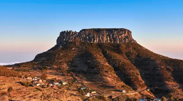 La imponente fortaleza de Chipude se alza sobre un paisaje rural con casas blancas y palmeras, bajo un cielo azul claro.
