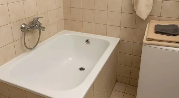 Biała wanna w łazience z beżowymi płytkami. Czy renowacja wanny to dobry pomysł? Obok pralka z ręcznikami.