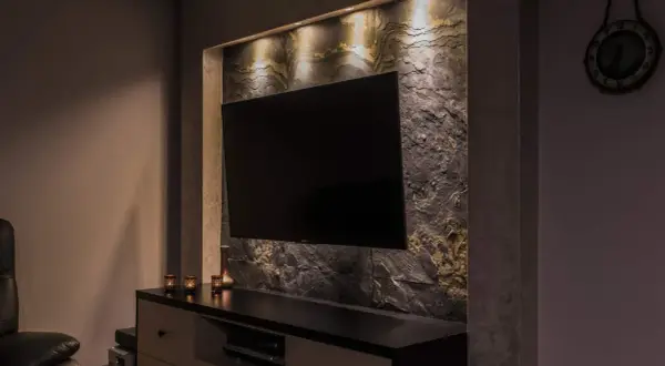 Nowoczesny salon z telewizorem wiszącym na ścianie wyłożonej kamieniem łupek. Oświetlenie podkreśla fakturę kamienia.