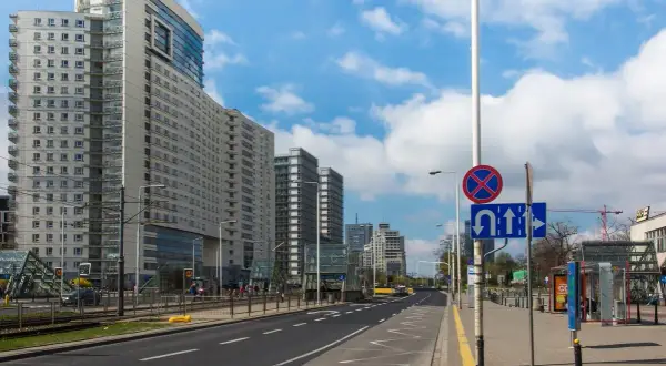 Ulica z nowoczesnymi budynkami i tramwajem. Na słupie widoczny znak D3 informujący o zakazie zatrzymywania się i dozwolonych kierunkach jazdy.