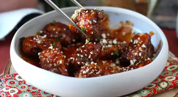 Chrupiący kurczak gochujang w sosie, posypany sezamem i orzeszkami, podany w białej misce.