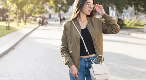 Młoda kobieta w stylowej kurtce wiosennej khaki, jeansach i z torebką na ramieniu spaceruje po parku.