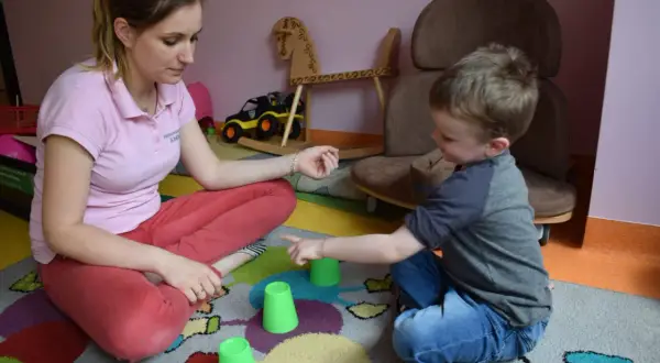 Terapia behawioralna autyzm: chłopiec i terapeutka bawią się kubeczkami na kolorowym dywanie.