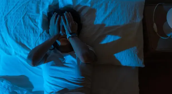 En person ligger i sängen, med händerna över ansiktet, i ett mörkt rum upplyst av blått ljus. En skugga av en hand syns på väggen, som om personen försöker vara fri från tvång.