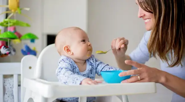 Mama karmi dziecko łyżeczką, realizując plan rozszerzania diety niemowlaka. Uśmiechnięta mama i maluch w krzesełku.