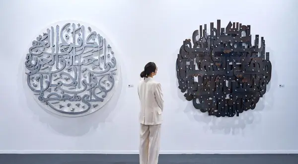 En kvinna i ljus kostym betraktar två runda konstverk med arabisk kalligrafi. Det vänstra är i metall, det högra i mörkare material. En atra daniel.