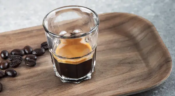 En liten shot med espresso, med crema, bredvid kaffebönor på en träbricka. En perfekt start på dagen, en stark espresso vs. vanligt kaffe.