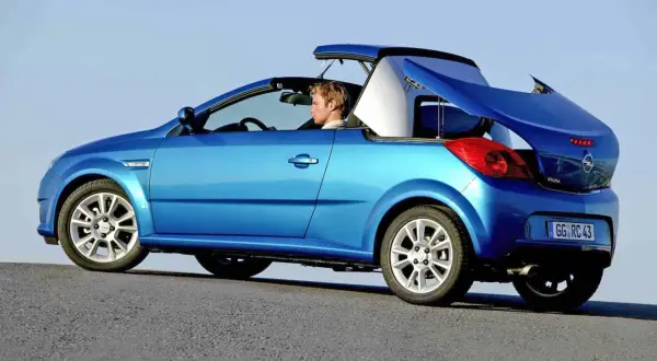 Niebieski kabriolet Opel Tigra z otwartym dachem, kierowca w środku, na tle błękitnego nieba.