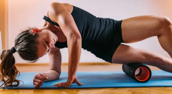 Woman performing a full body foam rolling routine on a blue mat, targeting her quadriceps.