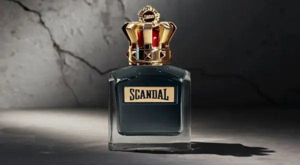 Elegancka butelka perfum Scandal męskie z koroną na nakrętce, stojąca na tle pękniętej ściany.