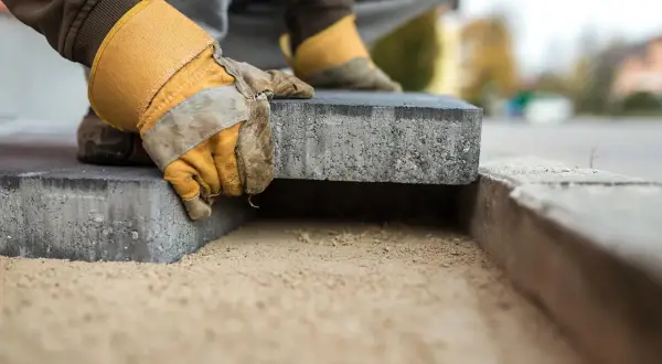 Mani guantate posano un blocco di cemento su un letto di sabbia, creando un nuovo marciapiede in pietra.